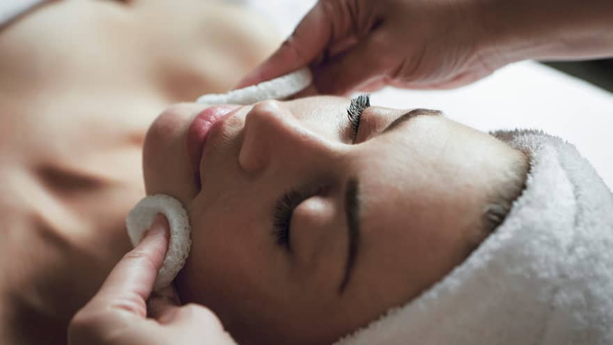 Woman with eyes closed, hair wrapped in towel, as spa attendant rubs cheeks with white pads