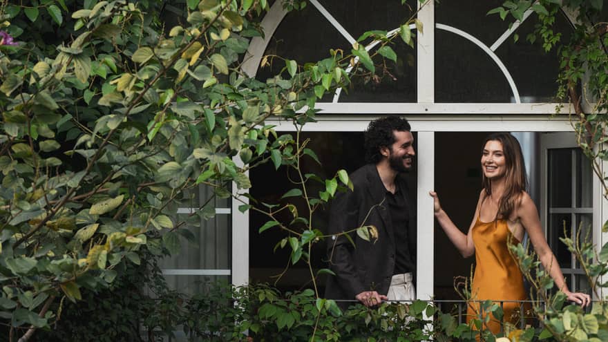Two people stand on a vine-covered balcony surrounded by foliage.