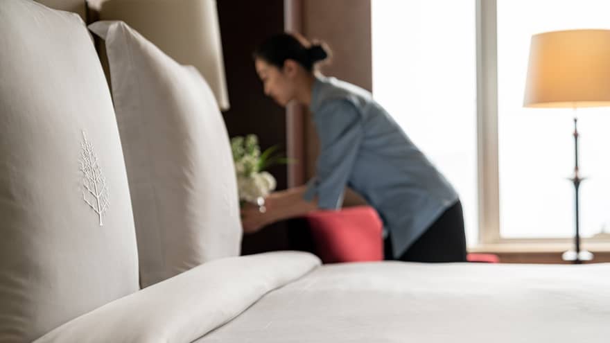 Close up of bed with white linens and FS logo, staff member arranging flowers in the background