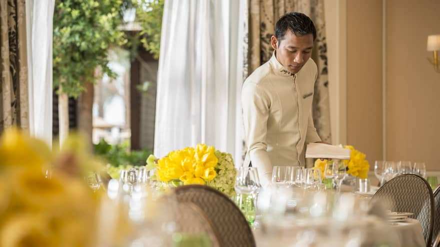 Hotel staff sets banquet table with wine glasses, yellow flowers in sunny event space