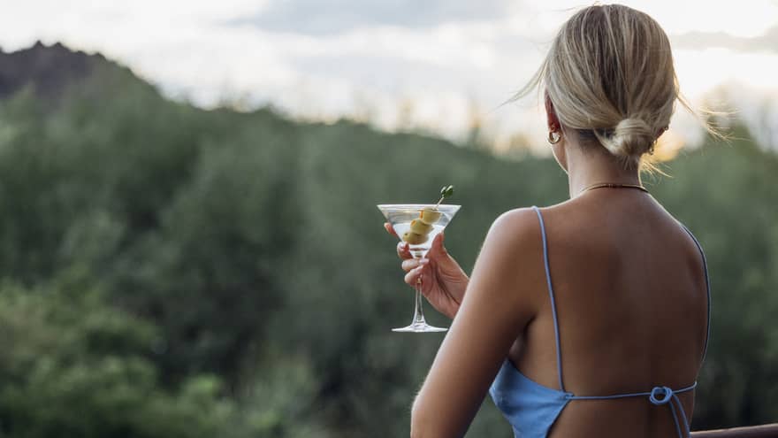 Guest standing on a balcony and holding a martini glass looking at the views of the forest and mountains.