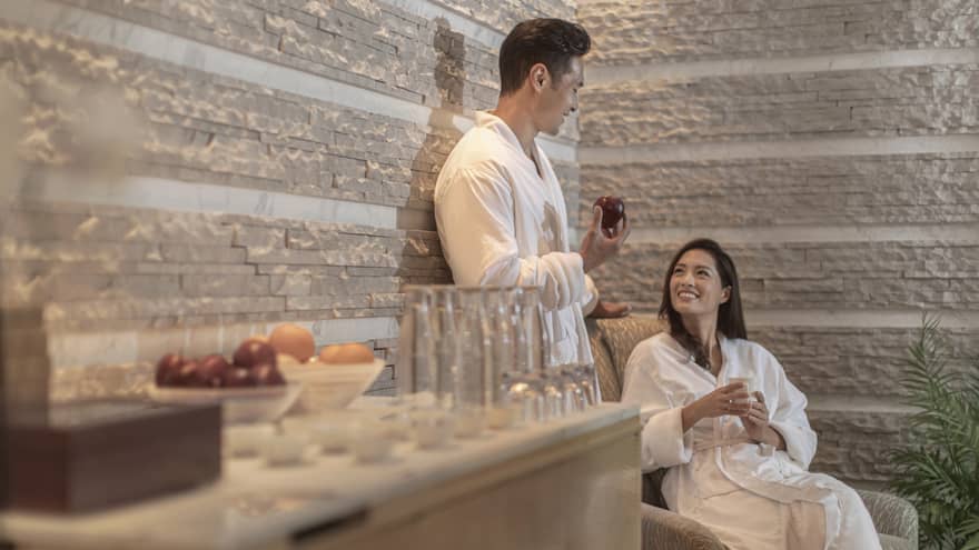 Two people in white robes relax in a spa lounge, one holding an apple. Various fruits and glasses sit on a wooden surface. The atmosphere is calm and inviting.