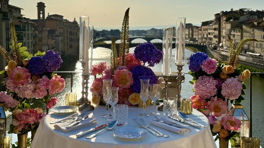 An elegantly set outdoor dining table overlooking a a river, with a sunny sky in the background.