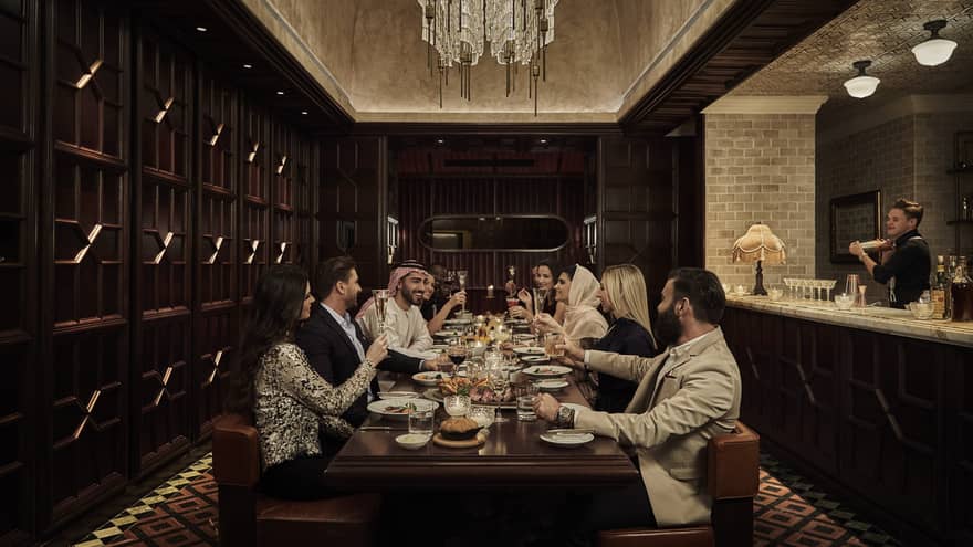 Ten people sit at a long, dark-wood rectangular table in a dark-wood panelled, speakeasy-style room with a crystal chandelier overhead and a marble bar counter and bartender to the side