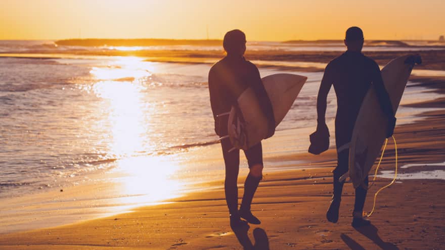 Silhouettes of two people carrying surfboards across beach at sunset