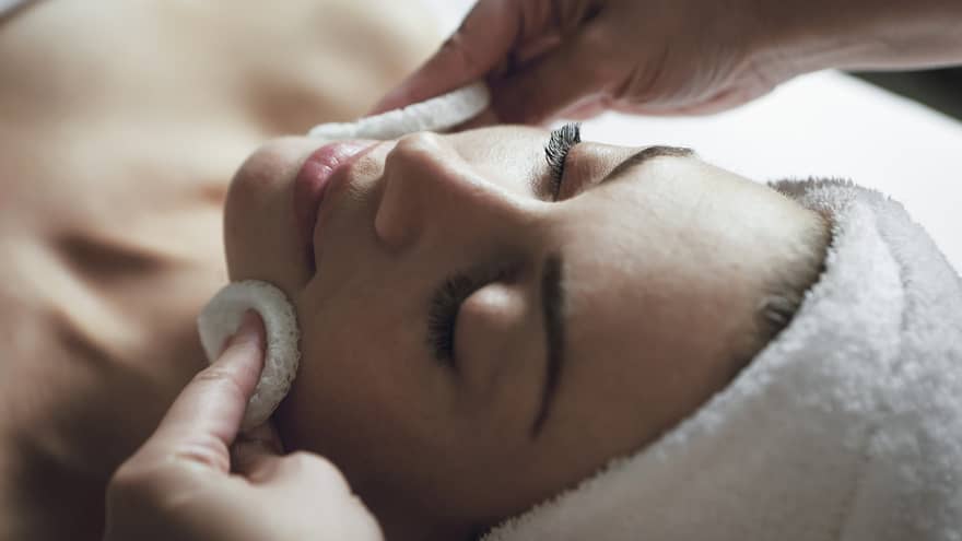 A detail of hands rubbing a sponge on a woman's face as she lies on a massage table with her eyes closed and hair wrapped up in a white towel
