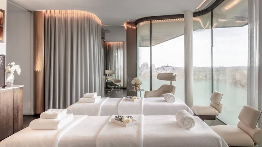 Elegant couples spa treatment room at the Four Seasons London Park Lane in Mayfair, offering a tranquil and luxurious setting for relaxation. The room features two side-by-side massage beds dressed in pristine white linens, surrounded by soft, floor-length curtains that ensure privacy and create a soothing atmosphere. Panoramic windows fill the space with natural light and provide a serene view of the skyline. Contemporary armchairs and tasteful decor elements like sculptures and floral arrangements enhance the spa's calming and exclusive ambiance.,Elegant couples' spa treatment room at Four Seasons London Park Lane in Mayfair, offering a tranquil and luxurious setting for relaxation. The room features two side-by-side massage beds dressed in pristine white linens, surrounded by soft, floor-length curtains that ensure privacy and create a soothing atmosphere. Panoramic windows fill the space with natural light and provide a serene view of the skyline. Contemporary armchairs and tasteful decor elements like sculptures and floral arrangements enhance the spa's calming and exclusive ambiance.