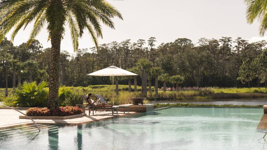 Outdoor swimming pool where woman lounges on patio chair under palm tree