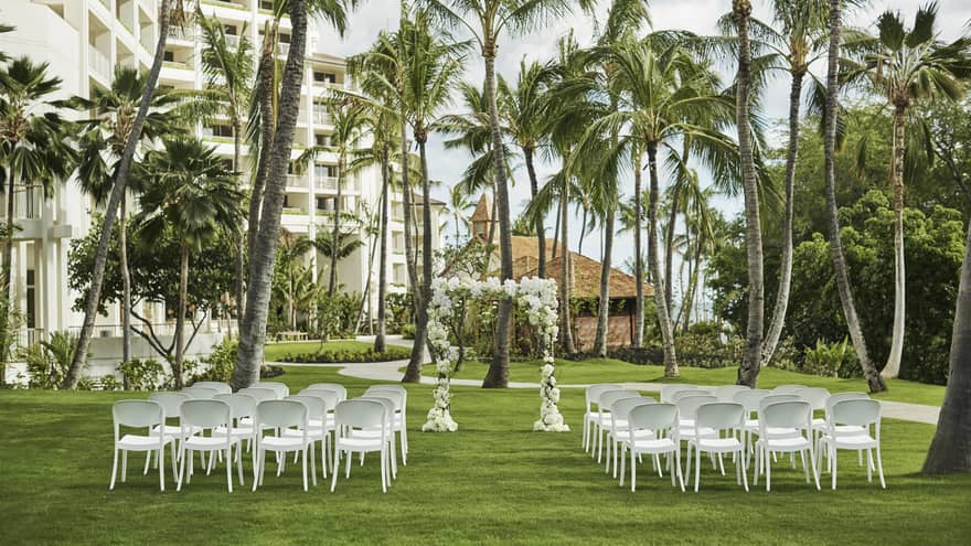 A picturesque outdoor wedding venue with elegant floral arrangements, white chairs on a lush green lawn, beneath palm trees