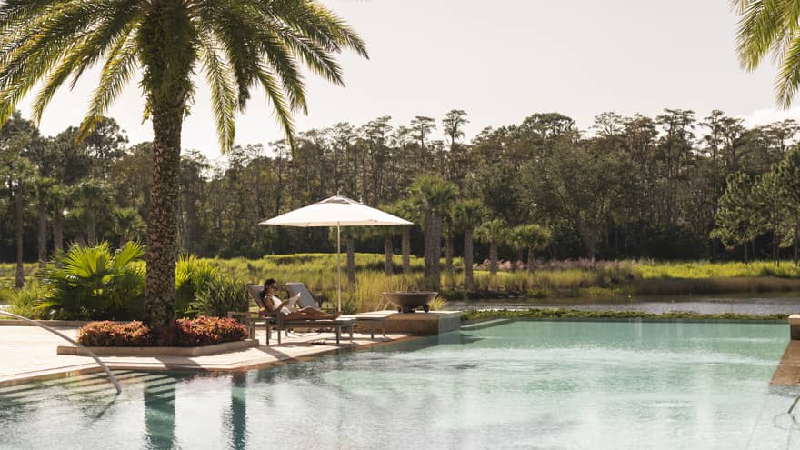Outdoor swimming pool where woman lounges on patio chair under palm tree