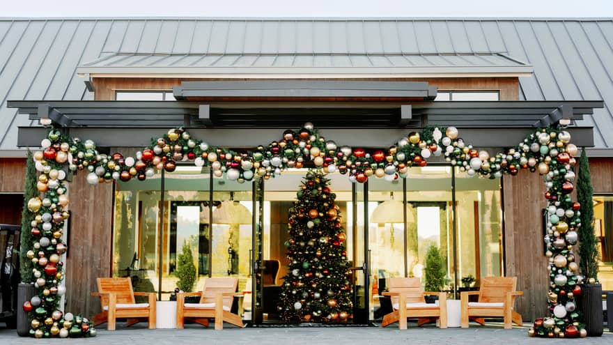Garland and colourful ornaments frame a glass-fronted entrance to a room with a large, colourfully decorated Christmas tree.