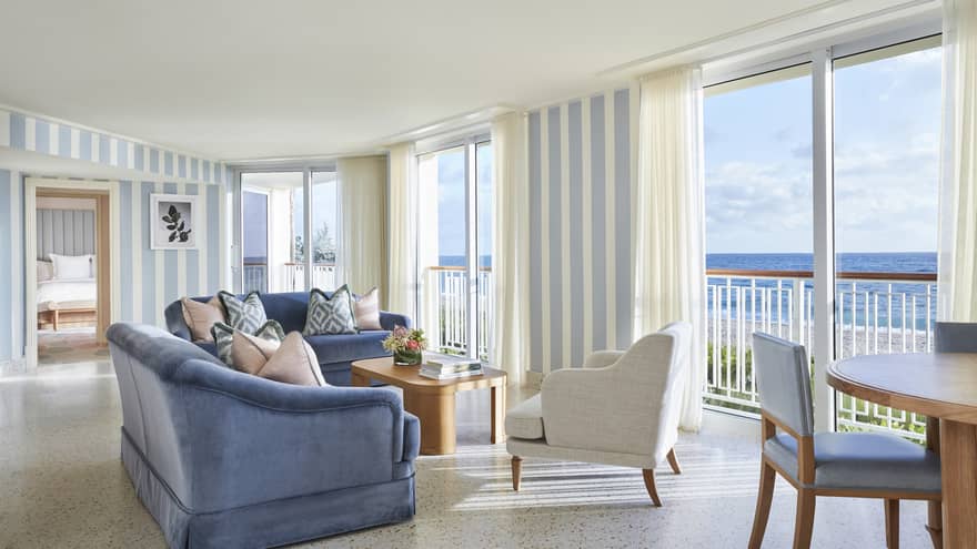 Living area in a suite, with blue sofas, a white armchair and floor-to-ceiling views of the ocean
