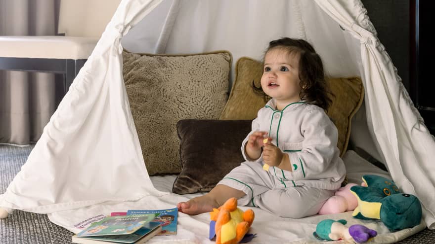 Toddler girl sits among toys and pillows in white play tent in room