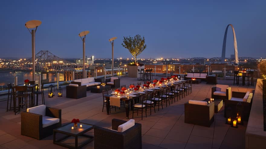 A panoramic view of the rooftop patio at night against the cityscape, featuring lounge chairs and long communal dining table with candles.