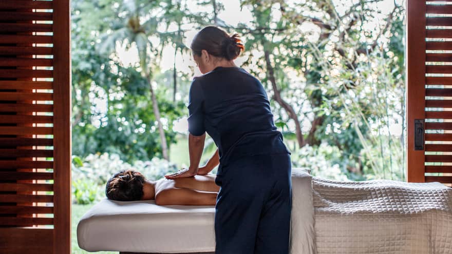 Spa staff massages woman's bare shoulders as she lies under sheet on massage table