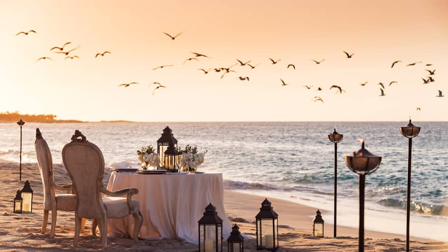 Romantic beachside dining setup with vintage chairs, a table decorated with flowers and lanterns and birds flying overhead