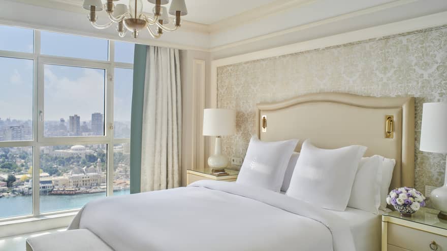Guest bedroom with king bed, white linens and cream tufted headboard; floor-to-ceiling window with views of the River Nile