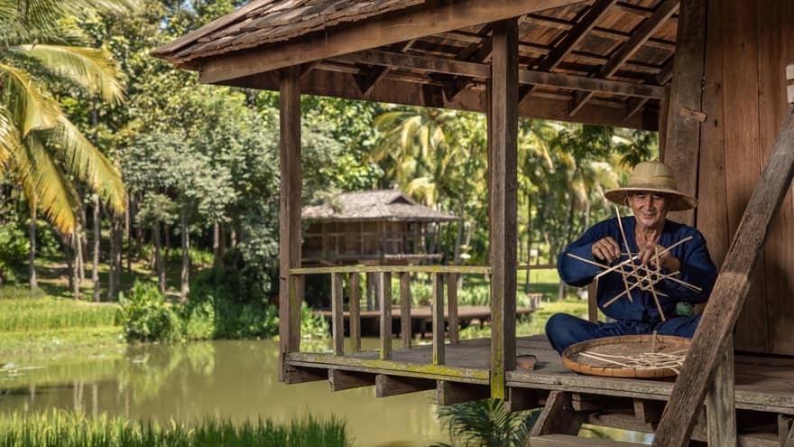 Man weaves a traditional Thai basket on the porch of a riverside pagoda