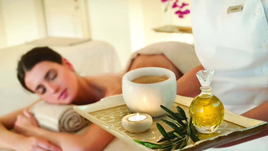Spa staff holds tray with tealight candle, perfume, marble bowl near woman lying on massage table
