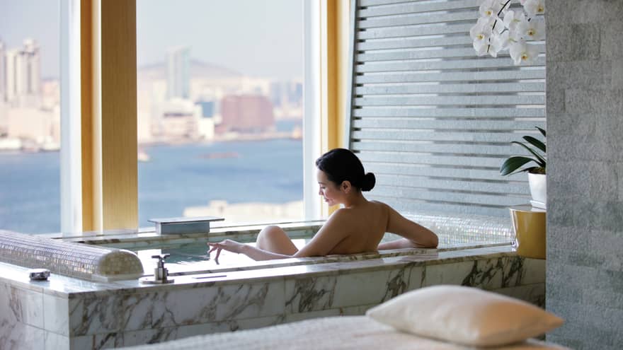 Woman's bare back above marble spa tub in tile bathroom by window with water, city views