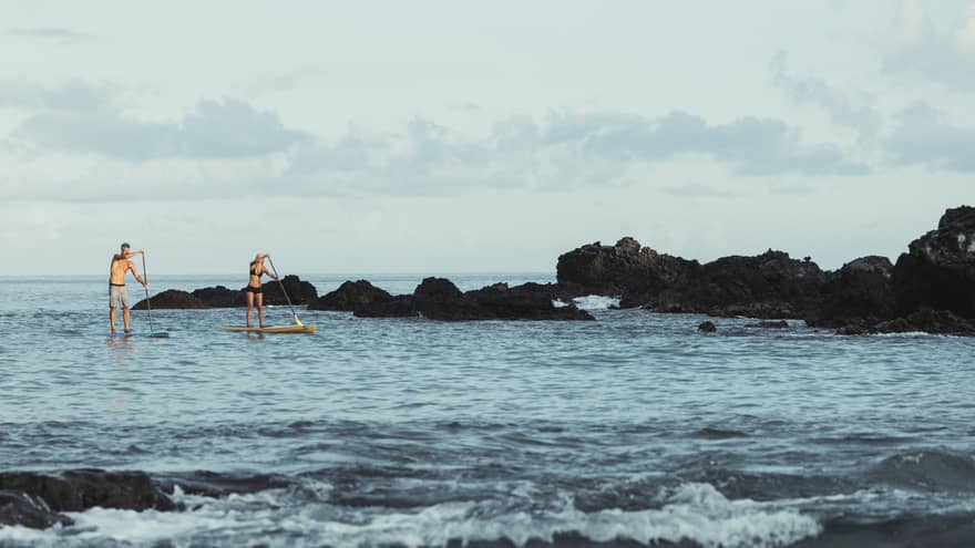 Two people balance on stand-up paddleboards in lagoon by black rocks