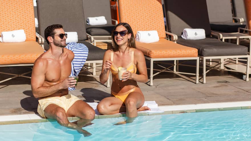 Man and woman sit on side of pool with feet in the water, and drinks in hand