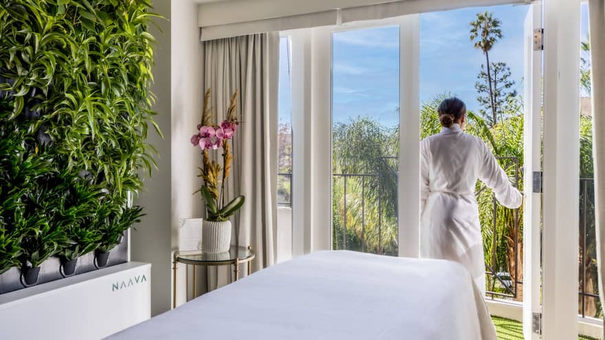 A woman walking outside from a massage room with large windowed doors and plants on one wall.