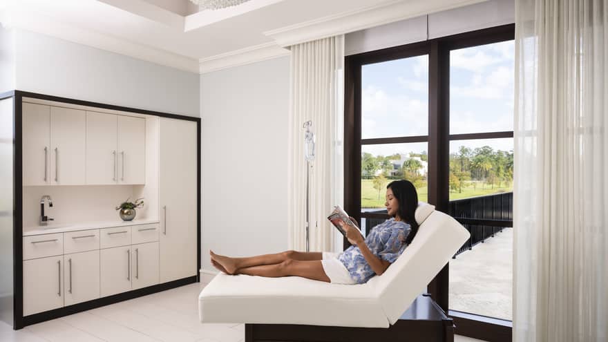 A woman reading a magazine on a folded mattress in a spa clinic with large window.