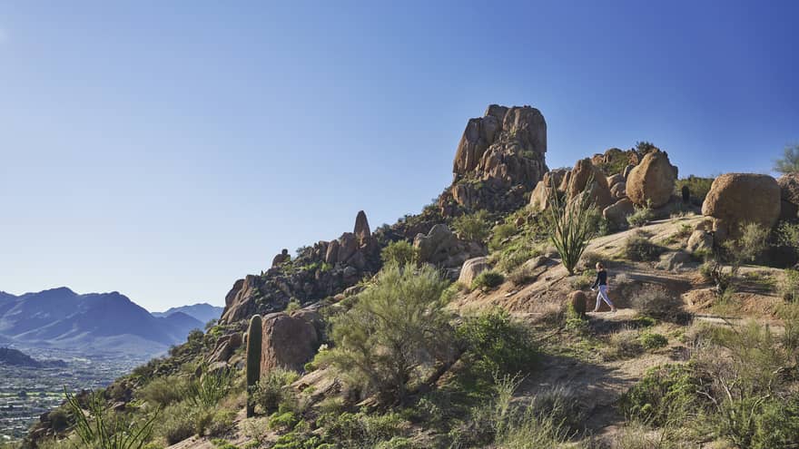 Woman walks along trail on rocky desert mountain under blue sky