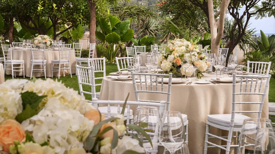 Outdoor wedding reception featuring white chairs and tables adorned with peach and ivory flowers in a garden setting