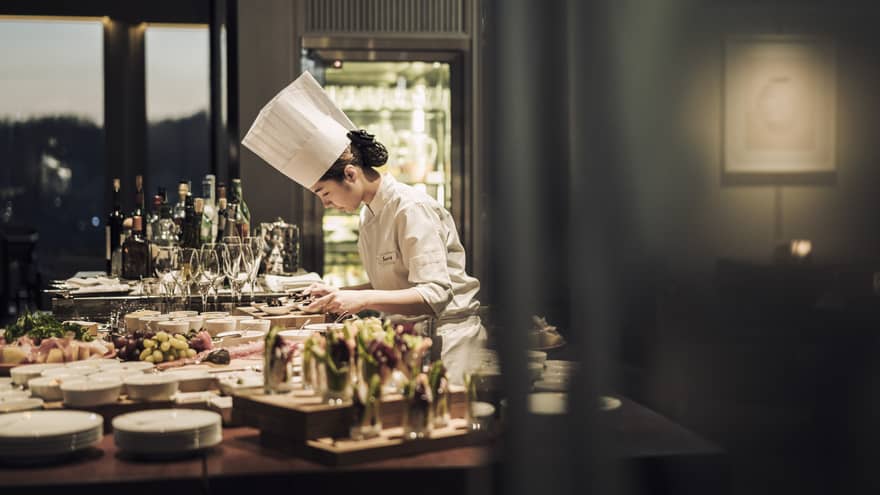 Chef wearing white uniform, hat stands garnishes appetizers on counter covered with dishes, meals