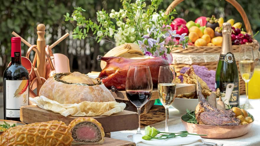 Close-up of food table with wine, basket with fruit, platters with roasted meats, fried duck, bread