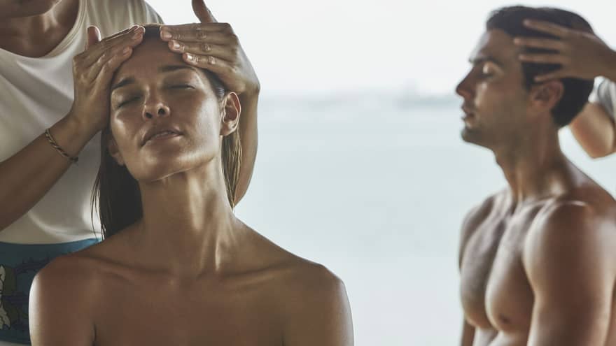 Close-up of woman and man with eyes closed as masseuses massage their temples