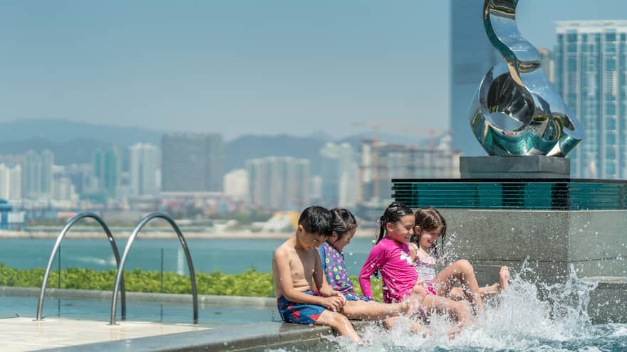 Four children sitting at the edge of a pool, splashing water with their feet. City buildings and mountains are visible in the background.