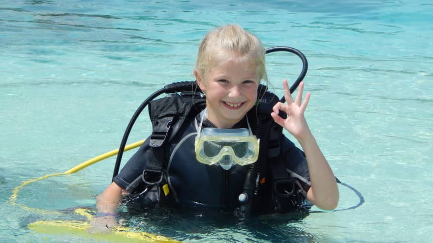 Young girl wearing scuba diving gear in water smiles, gives hand signal