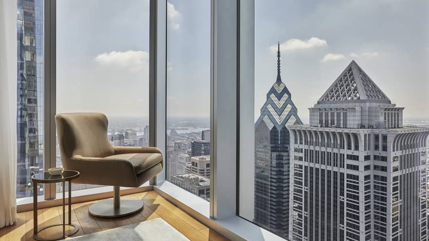 The corner of a hotel suite with large windows and a chair.