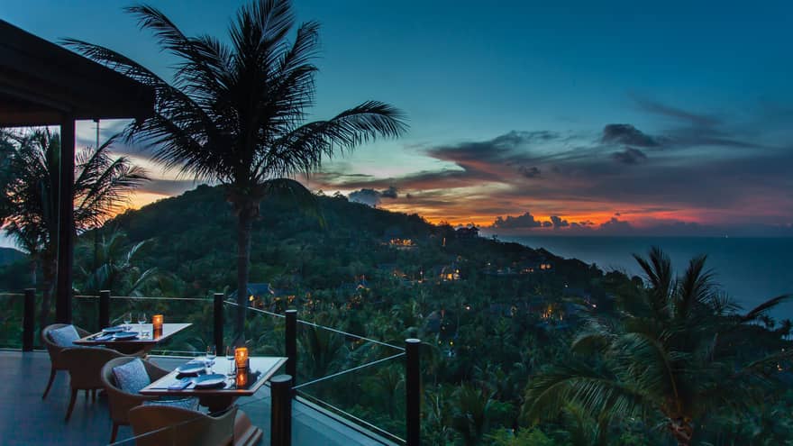 Corner of extended balcony with dining tables and chairs at dusk, expanse of forest, palm trees
