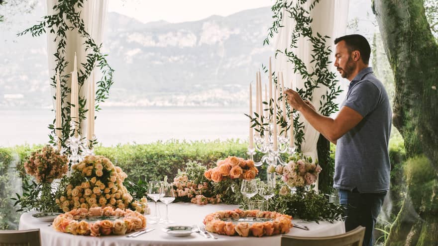 Man arranges tall candles in candelabra at table with pink rose floral arrangements, water views
