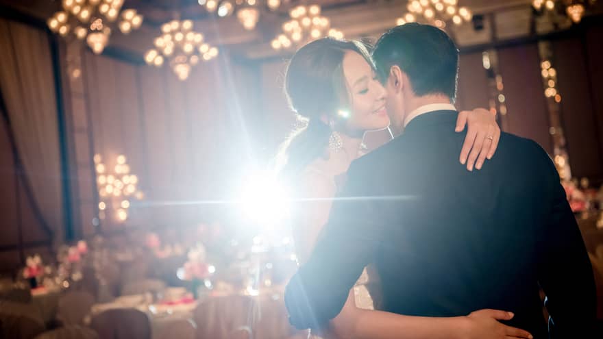 A bride and groom embrace each other in an indoor reception hall decorated with chandeliers and banquet tables