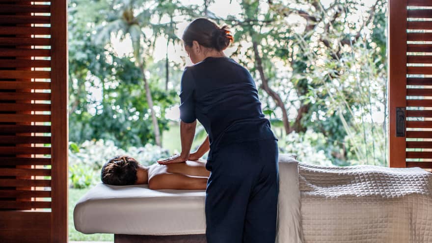 Spa staff massages woman's bare shoulders as she lies under sheet on massage table