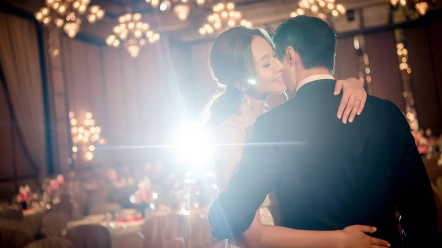 A bride and groom embrace each other in an indoor reception hall decorated with chandeliers and banquet tables