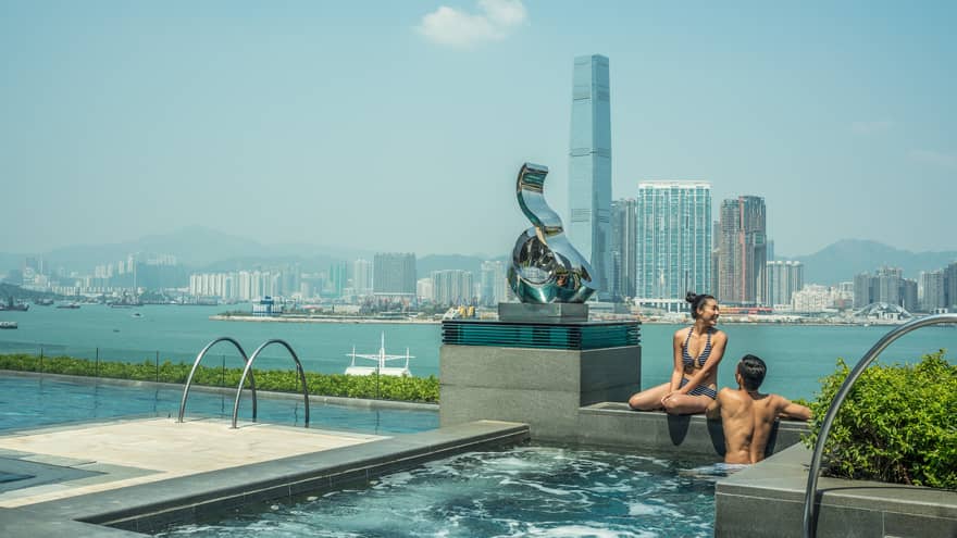 A couple in swimwear sit on the edge of an infinity pool with a harbor view, overlooking a city skyline, modern sculpture and mountains under a clear sky.