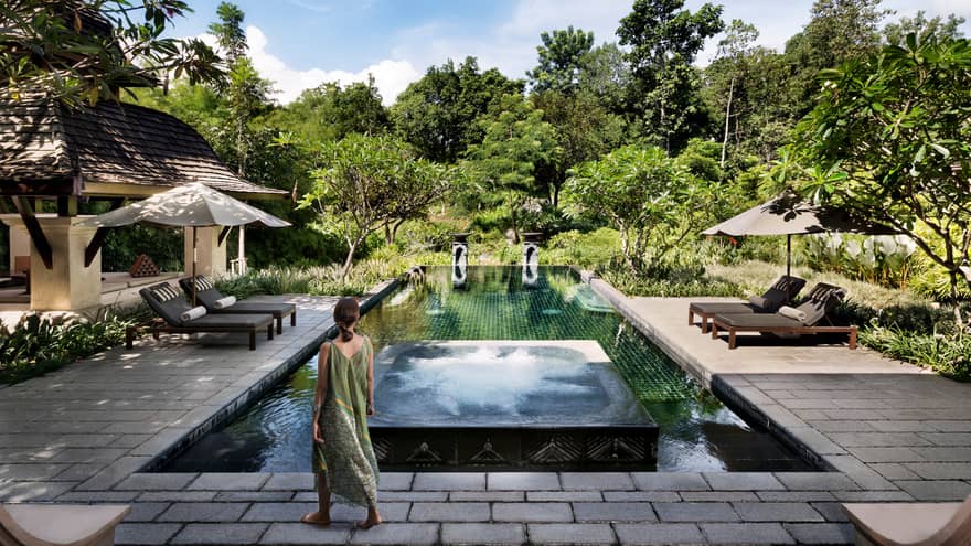 Back of woman in long beach dress walking towards residence swimming pool surrounded by tropical forest