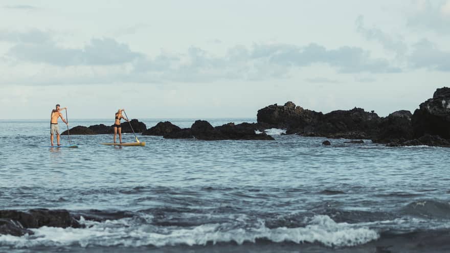 Two people balance on stand-up paddleboards in lagoon by black rocks
