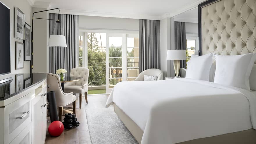 A hotel room with large open window, bed with white sheets, dresser, two chairs, an exercise ball and weights.