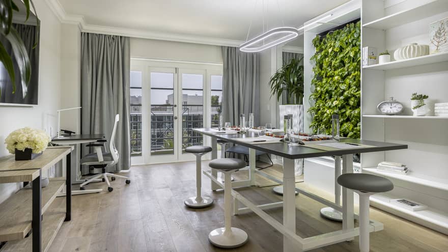 A brightly lit office with a large table, stools, a desk, shelves, and plants on the wall.