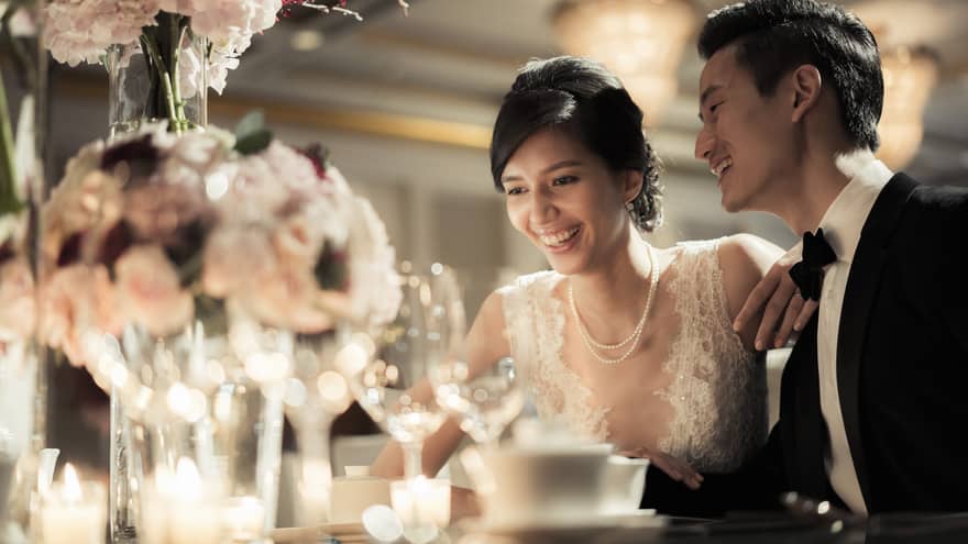 Smiling bride and groom at wedding dining table with glowing candles, pink roses