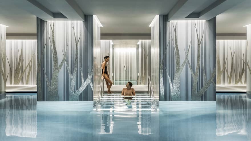 Woman in black swimsuit enters serenity pool while man sits on steps between blue and silver columns