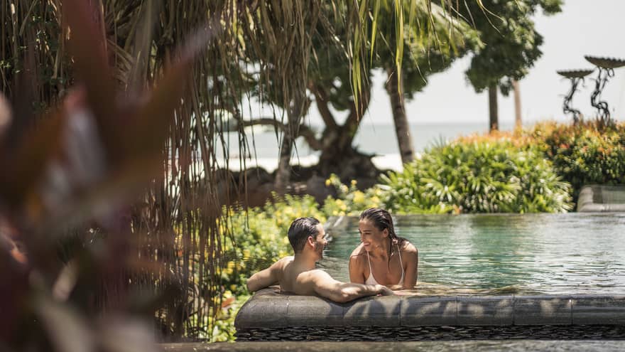 Smiling couple wades in outdoor swimming pool surrounded by tropical gardens