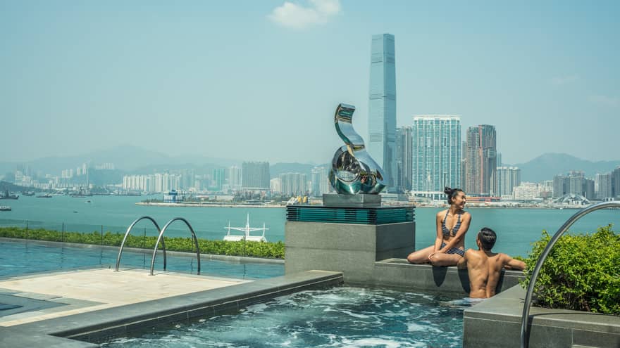 A couple in swimwear sit on the edge of an infinity pool with a harbor view, overlooking a city skyline, modern sculpture and mountains under a clear sky.